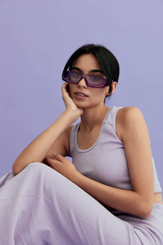 Young woman wearing purple sunglasses and a lilac ribbed top poses against a pastel lavender background, showcasing modern summer fashion and relaxed mood.