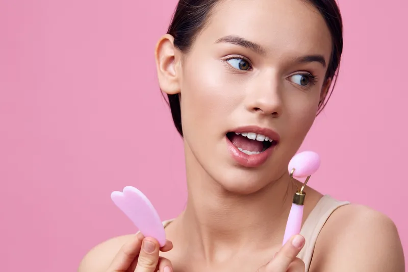 Young woman showcasing beauty tools against a pink backdrop, expressing excitement and curiosity about skincare techniques