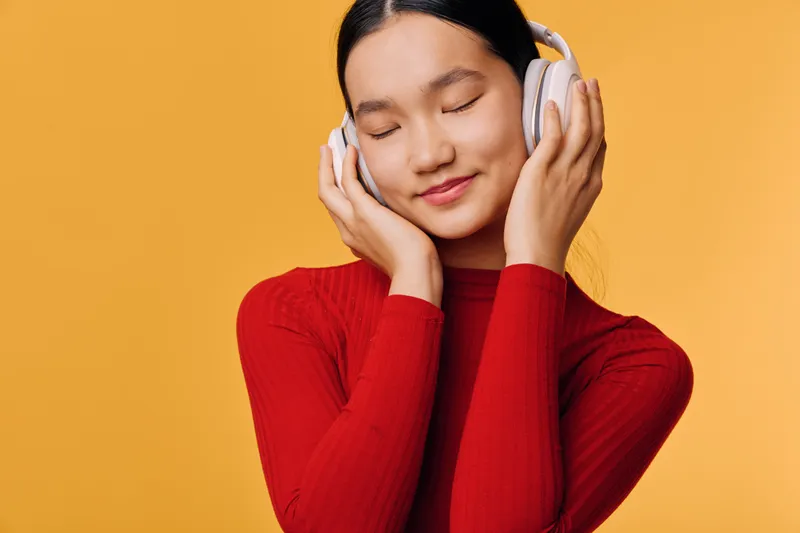 Young woman with closed eyes wearing headphones and red sweater enjoying music on orange background. Relaxation, technology, lifestyle and leisure concept with happiness and calm expression.