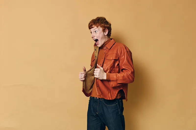 Young man with red hair wearing orange jacket holding brown hat with surprised expression on face standing against plain beige background in casual style and posture.