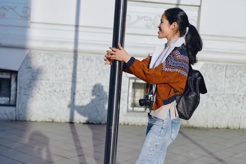 Woman wearing a brown jacket and jeans walks along a city street, smiling, holding a vertical pole with a backpack and camera strap visible, capturing an urban lifestyle moment