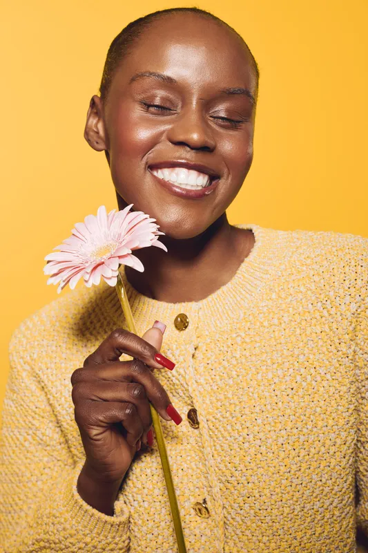 Smiling dark skinned African woman in yellow sweater holds pink flower close to face with closed eyes. Studio portrait on yellow background. Concept of happiness, beauty, and natural skincare.