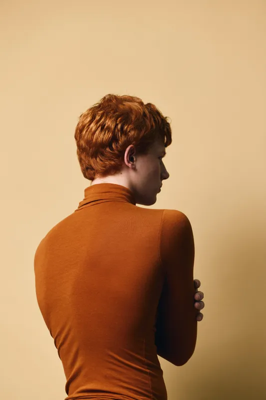 Redhead woman standing with back turned wearing brown turtleneck sweater on beige background, posing in thoughtful or introspective mood with minimalist style and warm tones.
