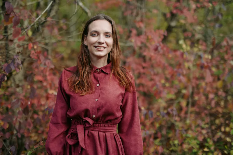 Portrait of a smiling young woman standing among autumn foliage, natural pose and candid expression conveying authenticity and a genuine, trustworthy presence in an outdoor lifestyle scene.