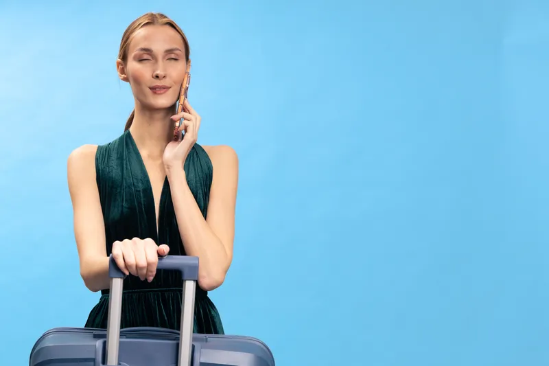 Young woman in green dress stands with suitcase and talks on phone against blue background. Concept of travel, communication, and summer vacation planning.