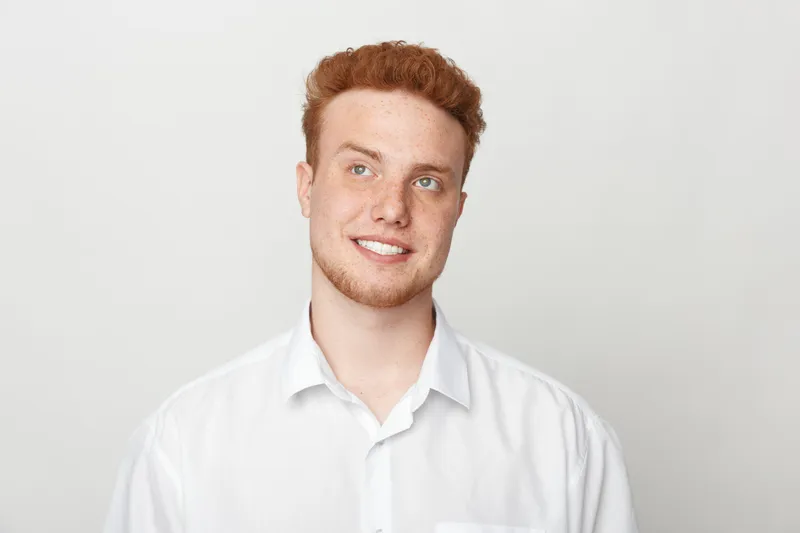 Smiling young man with red hair and freckles wears white shirt against light background. Portrait of confident male model with casual style and natural expression in studio lighting.