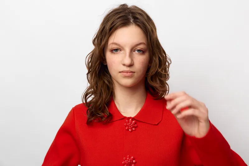 Young woman with wavy brown hair wears red sweater with floral buttons, posing against white background. Concept of casual fashion, portrait, and natural beauty with confident expression.