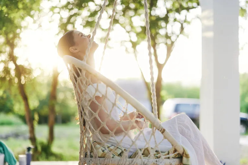 Serene young woman enjoying fresh air while relaxing on a swing in an outdoor setting surrounded by greenery and warm sunlight, embodying tranquility and peace