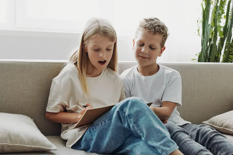 Two children sitting on sofa drawing together, focused on creative activity in bright room. Concept of childhood, creativity, learning, friendship, and home comfort.