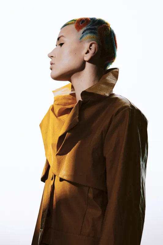 Young woman with colorful braided hair wearing a stylish brown jacket poses in profile against bright white background, showcasing modern fashion and artistic hairstyle.