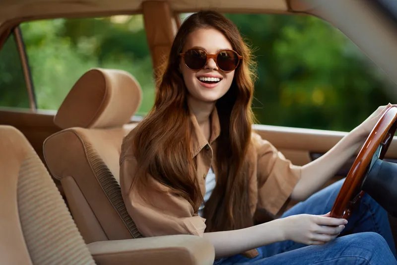 Happy young woman with long hair driving a vintage car, wearing sunglasses, in a vibrant green outdoor setting, evoking a sense of freedom and adventure.