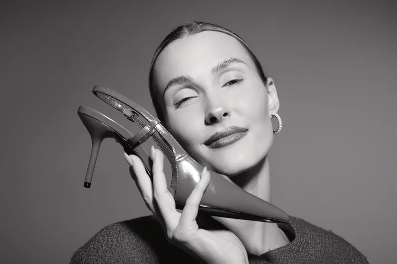 Closeup of smiling white woman with fair skin holding high heel shoe near face, studio portrait on plain background. Concept of fashion and elegance.