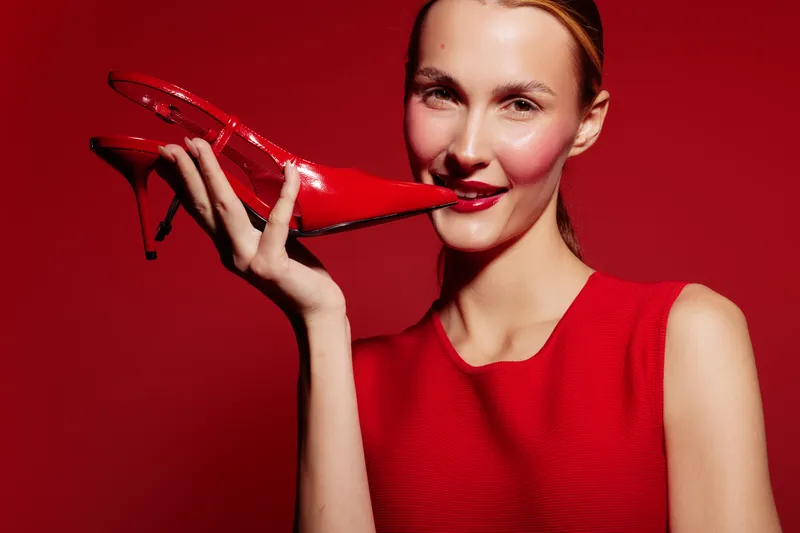 Young woman in red dress holds red high heel shoe near face, smiling confidently against red background. Concept of fashion, style, elegance, and beauty.
