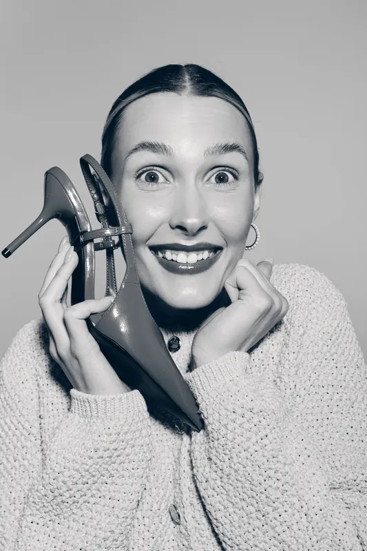 Excited white woman with fair skin holds high heel shoe near face, smiling with wide eyes. Concept of fashion, style, happiness, and shopping.