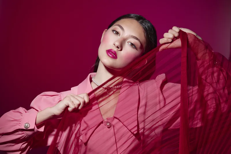 Young woman with glossy lips and smooth skin posing with a translucent red fabric against dark background. Creative beauty and fashion concept with vibrant colors and dramatic lighting.