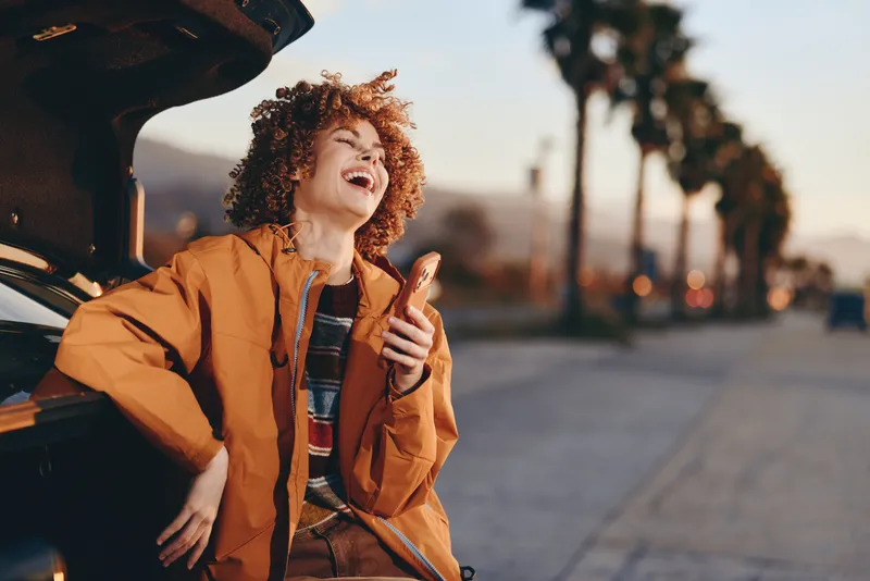 Woman lifestyle by car, smiling with curly hair in a warm sunset street, wearing casual jacket, open door scene, palm trees in background, joyful travel mood happy laughter