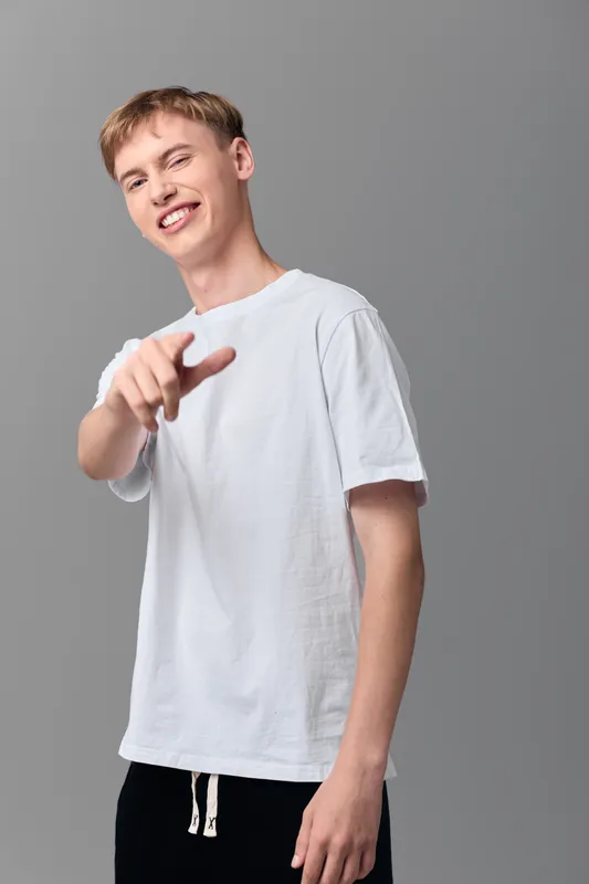 Smiling man pointing at camera in a casual white tee and black shorts against a grey studio backdrop, conveying friendliness, approachability, and a clear invitation to action for marketing and