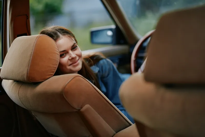 Young woman smiling in a vintage car, showcasing a relaxed and joyful expression in a soft natural setting, perfect for lifestyle and travel themes.