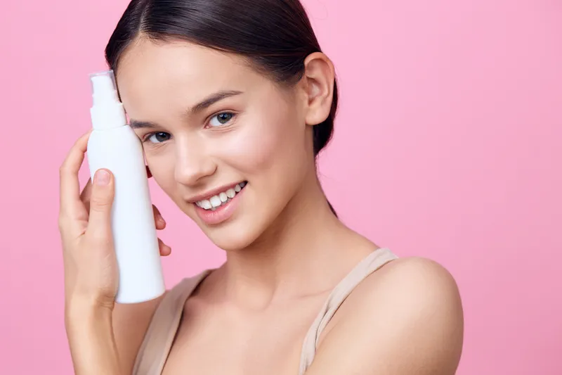 Young woman with clear skin holding a skincare bottle on a pink background, promoting beauty and self care products, showcasing radiant beauty and confidence