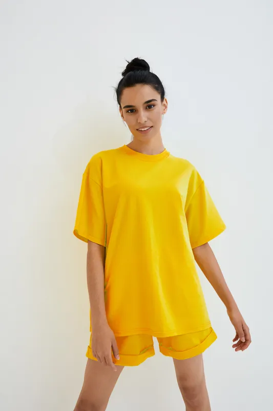 young woman wearing bright yellow casual oversized t-shirt and shorts standing against white wall with relaxed smile and natural pose in minimalistic studio setting