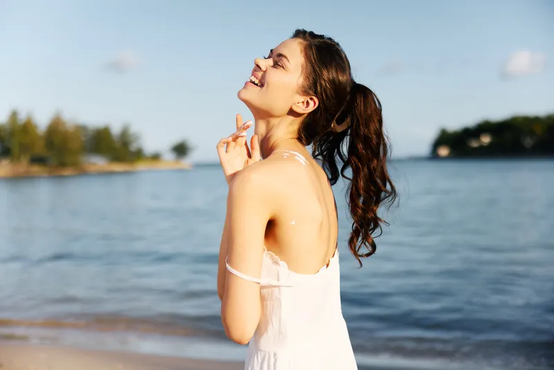 Joyful woman stands on a sunny beach, smiling with eyes closed, wearing a white sundress as gentle waves lap the shore, capturing carefree summer mood.