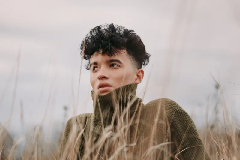 Close-up portrait of a young person in tall grass, captured with a natural, authentic expression and realistic tones, emphasizing genuine emotion and an unposed documentary style.