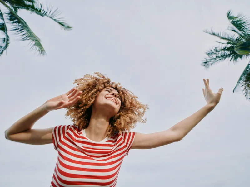 Joyful young woman in a red striped tee stretches arms toward a bright sky, framed by palm trees, capturing carefree summer energy and a vibrant mood of freedom and happiness.