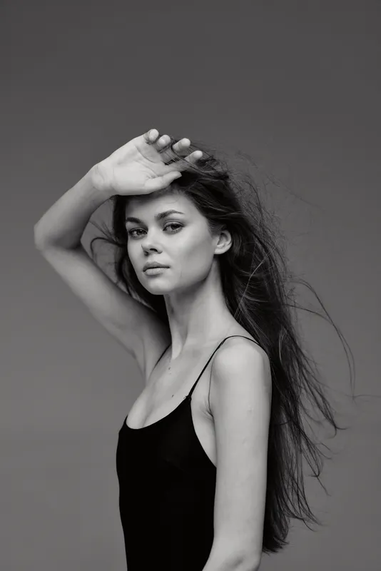 Black and white portrait of a young woman with long hair wearing a black top, posing with her hand raised in natural light.