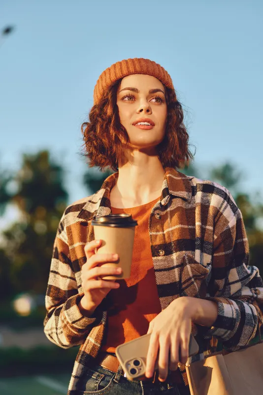 Woman coffee phone beanie plaid portrait outdoors smile. Candid lifestyle portrait of a young woman holding a takeaway cup and phone, golden hour glow, authenticity and mindful living mood.