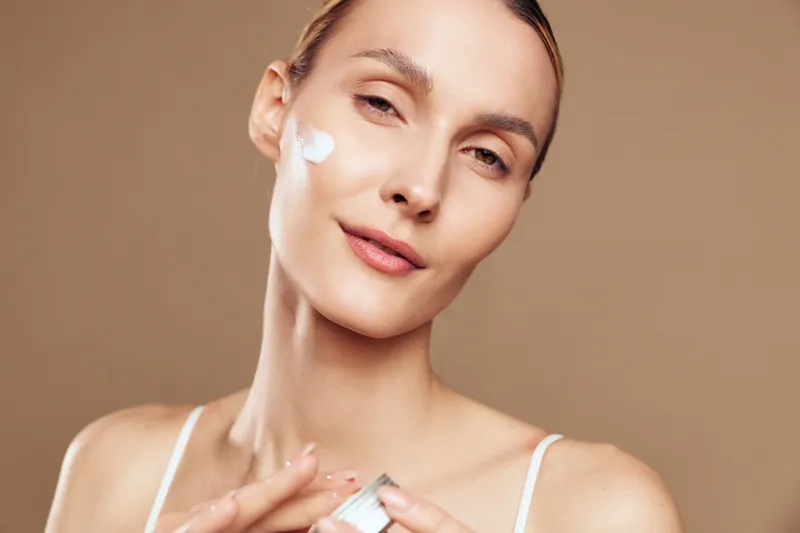 A young woman applies white face cream on her cheek in a close-up portrait with a soft brown background. The skincare concept emphasizes natural beauty and self-care routines.