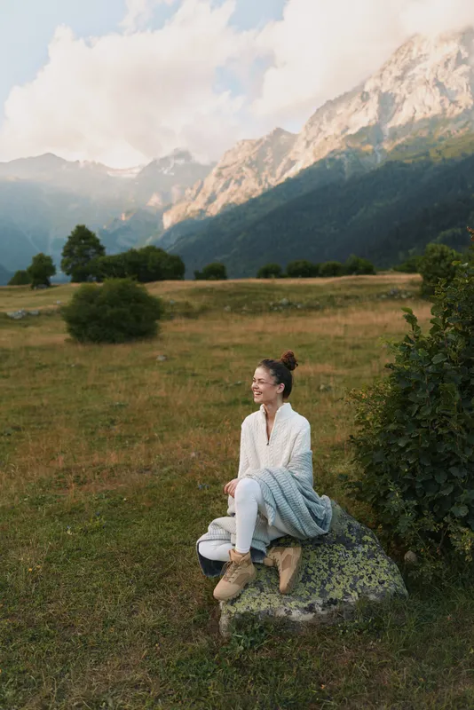 Young woman with brunette hair sits on a rock in a mountain meadow enjoying a peaceful moment surrounded by nature and distant peaks under a partly cloudy sky.
