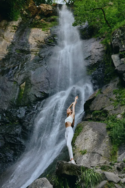 woman stretching near waterfall in nature, wearing white sportswear, enjoying outdoor fitness and calm environment, healthy lifestyle concept