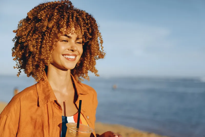 Woman with curly hair smiling in an orange jacket, holding a drink with straw by the seaside, ocean horizon in bokeh, summer travel portrait, outdoor daylight