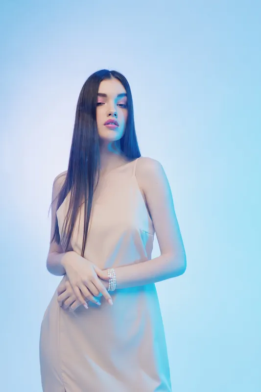 Young woman with long straight hair poses in a beige dress against a blue background. Concept of beauty, fashion, elegance, and modern style.