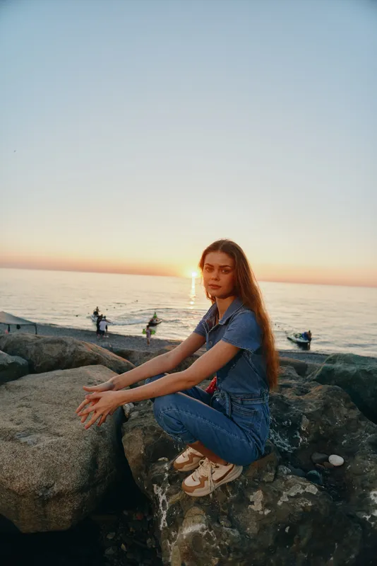 young woman sitting on rocks by the sea during sunset wearing casual denim clothes with calm ocean background and warm evening light