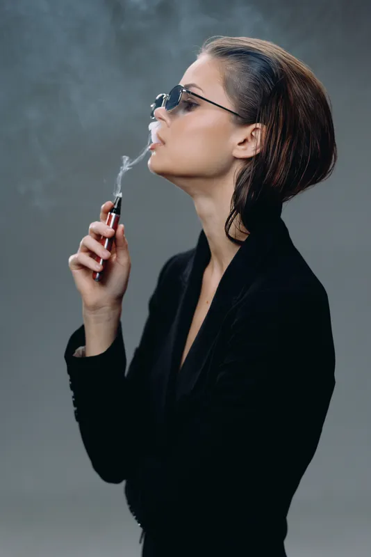 Stylish woman smoking electronic cigarette in dark outfit and sunglasses, blowing vapor smoke against gray background.