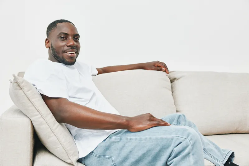 Relaxed young African American man in casual white t shirt sitting on a light colored sofa, smiling, conveying comfort and ease