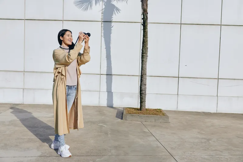 Camera in hand, woman wears trench coat, outdoor street scene by a white wall with palm shadow, candid fashion moment captured in urban photography and mood