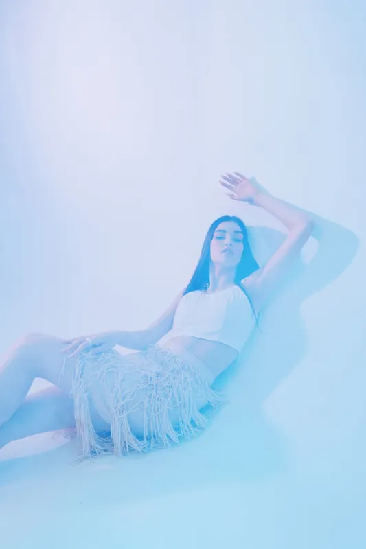 Young woman in fashionable white top and fringed skirt poses against soft blue background. Concept of beauty, fashion, style, and modern elegance in studio lighting.