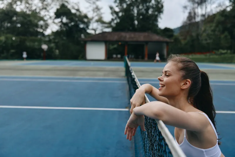 Smiling young model enjoying sunny day on tennis court, wearing sporty attire, expressing joy amidst greenery and blue tones