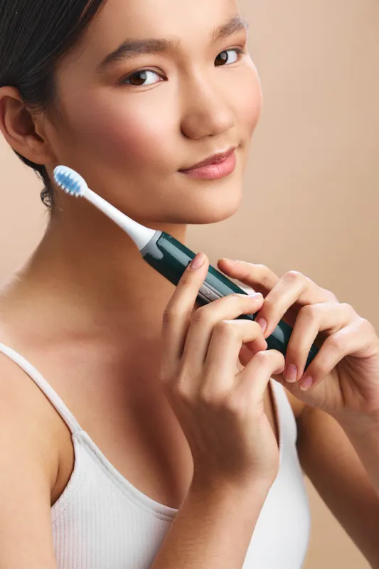 Asian woman holds a toothbrush close to her face, smiling softly as she freshens her routine in a clean bathroom. The scene conveys healthy habits, personal care, and calm confidence in a modern