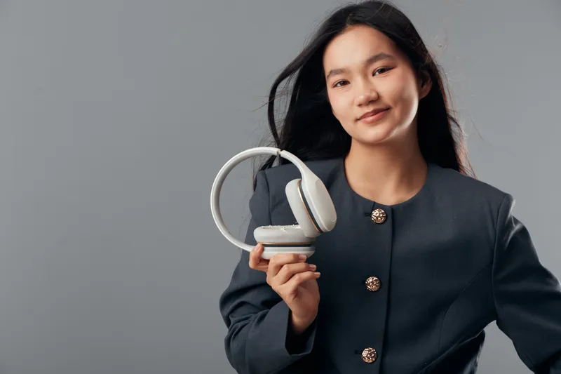 Young woman wearing dark jacket holds white headphones in hand smiling, portrait with long black hair in studio. Female person, fashion, technology and audio equipment on gray background.