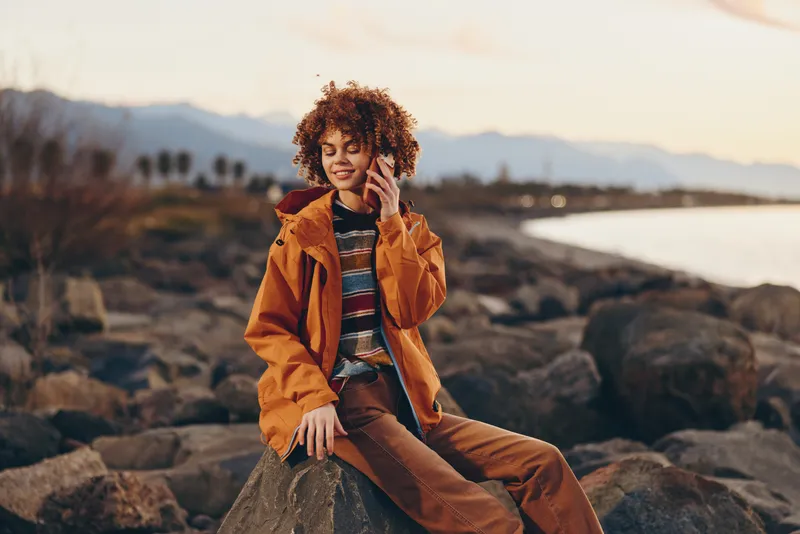 Woman lifestyle portrait, curly hair, orange jacket, outdoors at sunset by rocky coast, she sits on stones, warm golden light, calm relaxed mood, travel nature