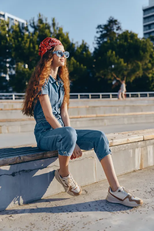 Lifestyle woman in modern boho-western denim street style sitting outdoors, wearing a red bandana and sunglasses in warm film color tones.
