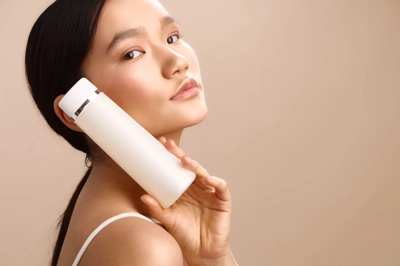 asian woman holds a skincare bottle in a soft studio setting, exuding calm confidence and care. her smooth skin and gentle smile convey beauty and wellness.