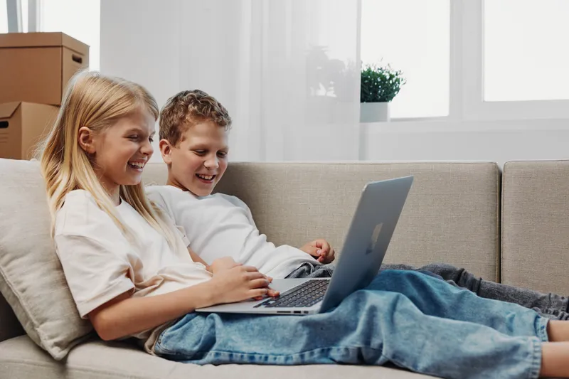 Happy children sitting on a sofa using a laptop, enjoying technology together in a bright living room. Concept of friendship, learning, and digital entertainment.