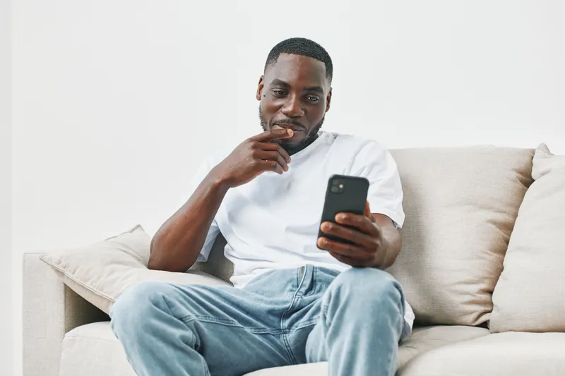 Smiling Black man sitting on a couch, looking at his smartphone, dressed casually in a white t shirt and blue jeans, relaxed home atmosphere