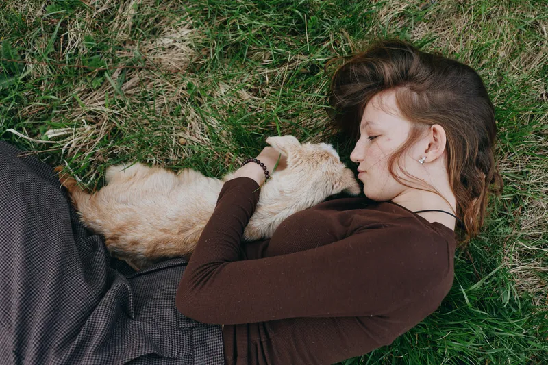 Woman lies on grass cuddling a dog, cozy outdoor moment in a brown sweater, natural light, relaxed lifestyle scene, affectionate bond, peaceful mood and quiet countryside feeling.