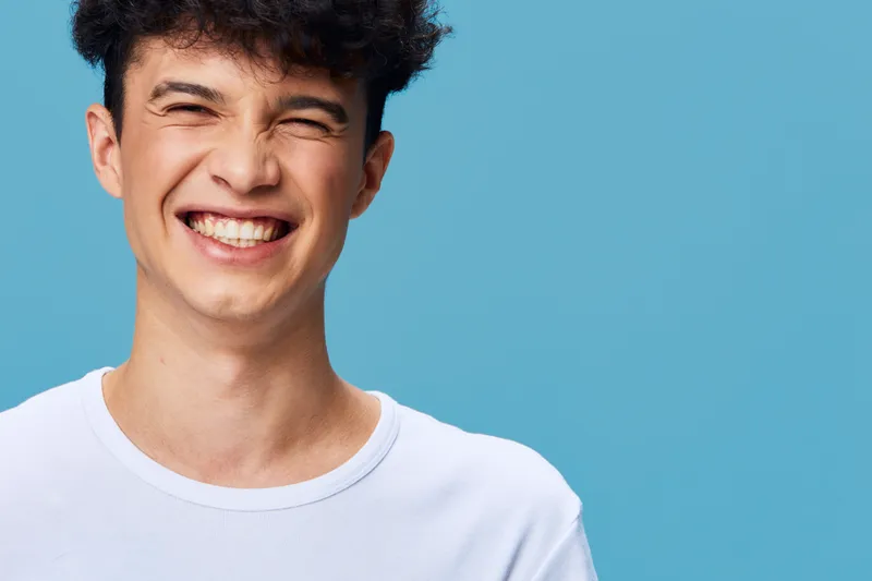 smiling man with curly hair wearing white shirt stands against blue background, expressing joy and happiness with bright facial expression and casual style.