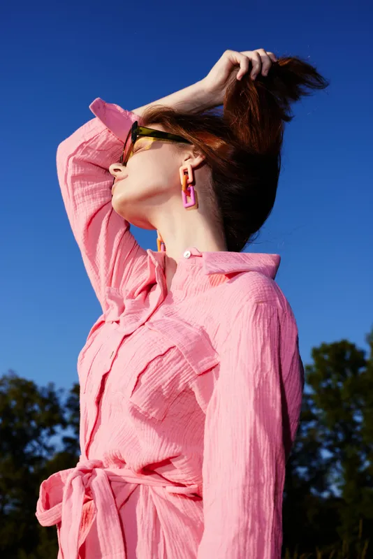 Stylish woman in a pink shirt dress posing against a clear blue sky, showcasing confidence and modern fashion in a vibrant and seasonal setting